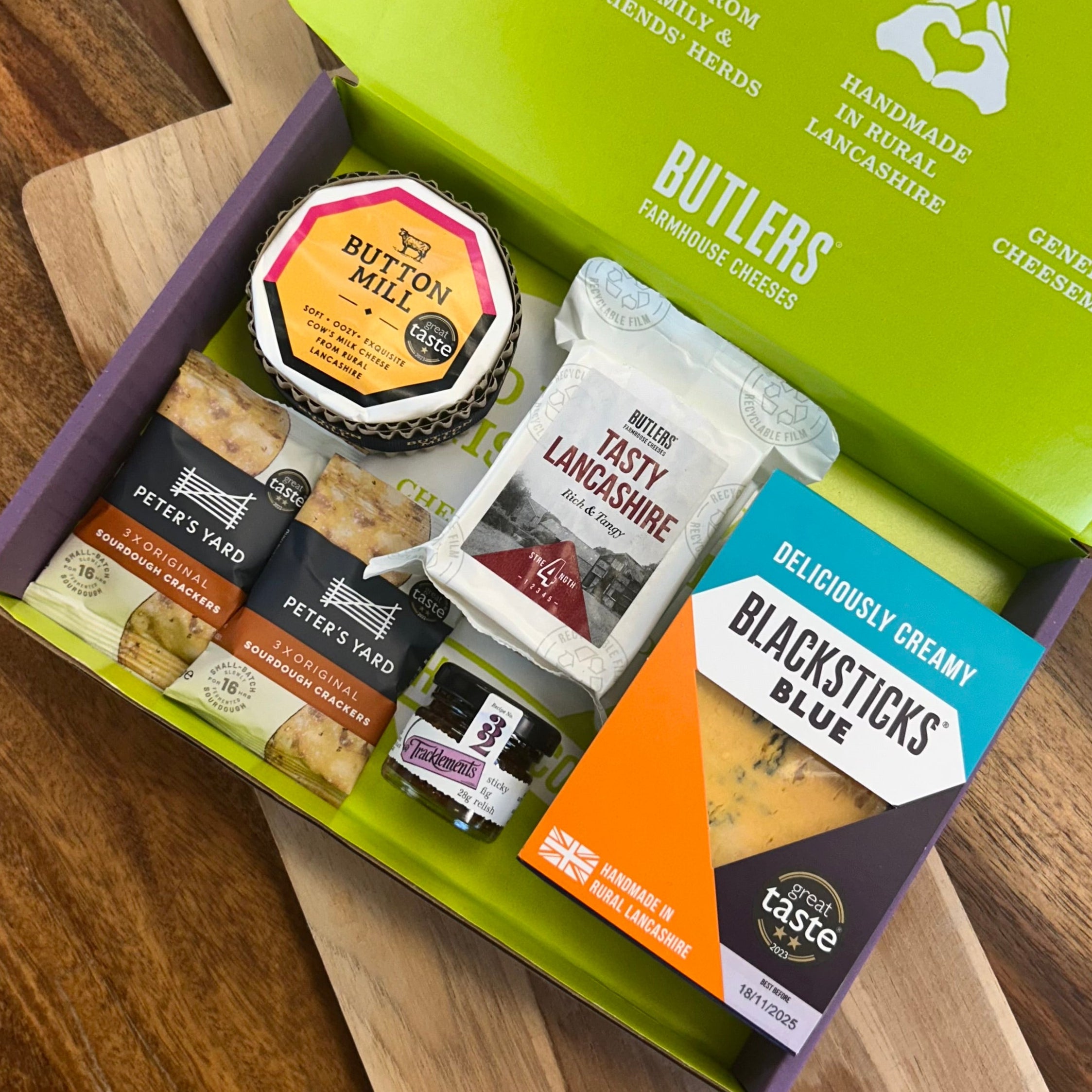 Butlers Farmhouse Cheeses' Perfect British Cheeseboard open on a wooden surface. Containing Blacksticks Blue, Tasty Lancashire and Button Mill, as well as crackers and relish.